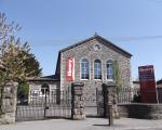 A picture of a grey stone building with three windows, an iron fence and a tall red sign