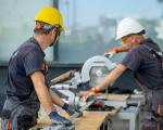 Two workers in hard hats cutting a metal beam with a circular saw