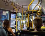 An image of people inside of a bus, seated and standing while holding onto the yellow support bars