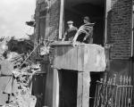 A black and white photo showing bomb damage caused to British houses with people talking and waving the Union Jack flag