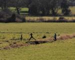 A child walks across a small bridge with their arms outstretched, while their family looks on. They are walking in a waterlogged field, with a village skyline visible in the background