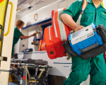 The back of an ambulance with door open. A paramedic is stepping out of the ambulance, carrying equipment. Another paramedic is inside the ambulance.