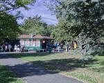 People sit outside and queue by a building with 'cafe' painted on it in red and green. Trees frame the image. 