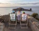 A photo showing two women sitting in traditional wooden deckchairs in Prince Consort Gardens overlooking Birnbeck Pier. A young girl with blonde hair stands between them. All three of them are wearing colourful party hats. Signs on the back of the deckchairs read £10m for Birnbeck Pier.