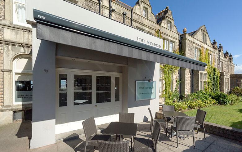 A white rectangular shop front attached to a Victorian building, with outside tables and chairs and a sign reading The Terrace