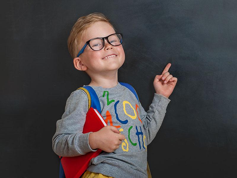 young blonde boy wearing a red backpack while smiling and pointing in the air.