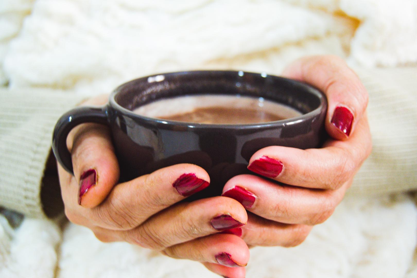 hands with red painted fingernails holding a warm drink in a black ceramic mug