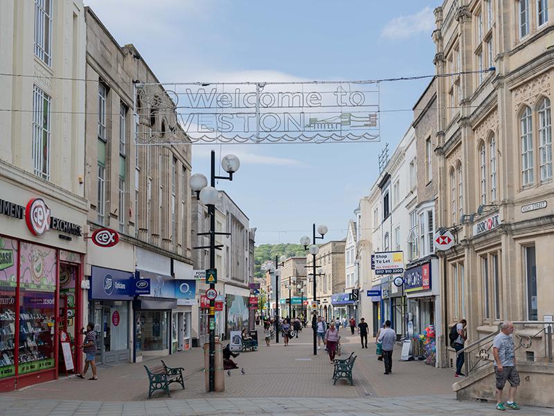 A picture of people milling around the old buildings on Weston High Street, with a Welcome to Weston sign overhead