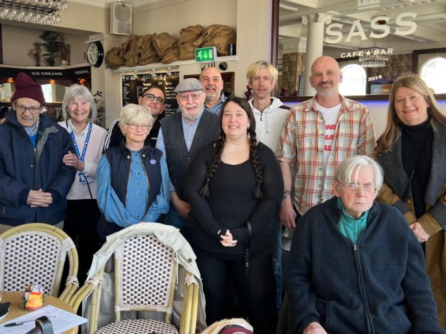 A group of men and women standing in a café smiling at the camera.