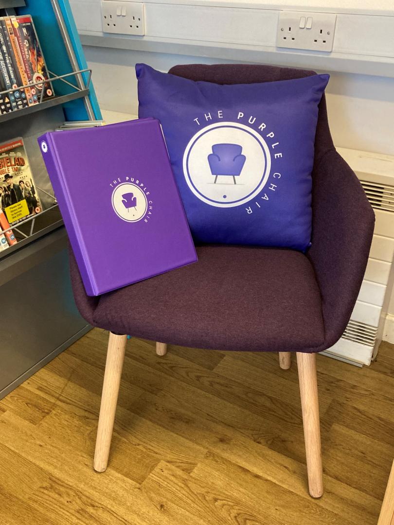 A photo of a purple chair in a library that has a purple cushion and purple ring binder folder on it. Both the cushion and folder have a logo on that says 'The Purple Chair' with a picture of a purple chair on it.