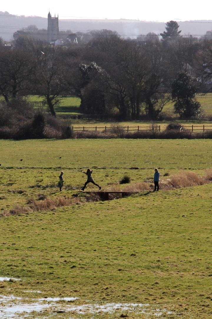 A child walks across a small bridge with their arms outstretched, while their family looks on. They are walking in a waterlogged field, with a village skyline visible in the background