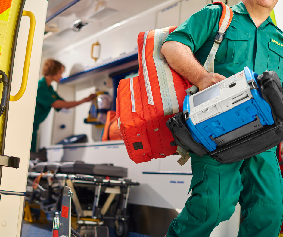 The back of an ambulance with door open. A paramedic is stepping out of the ambulance, carrying equipment. Another paramedic is inside the ambulance.