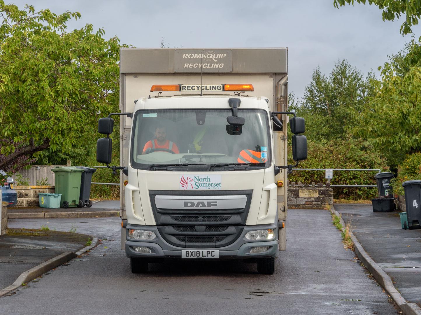 A recycling and waste vehicle drives down the middle of a residential street lined with trees. A man wearing a high-vis jacket is sitting in the driver's seat