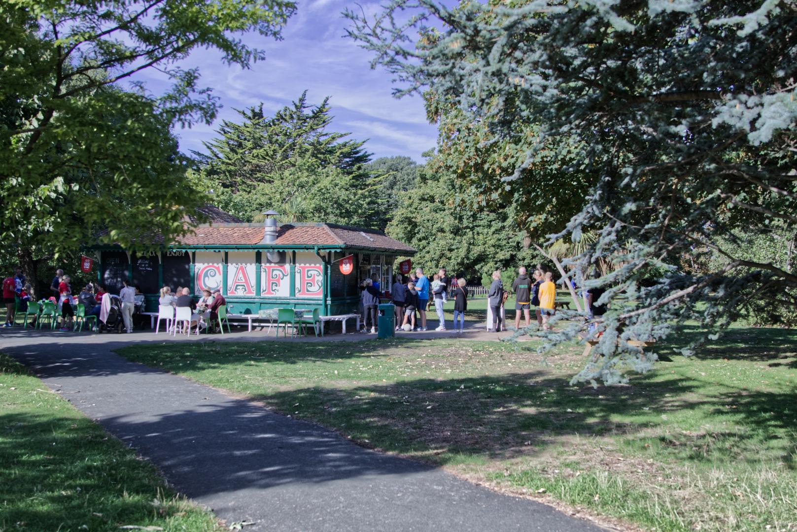 People sit outside and queue by a building with 'cafe' painted on it in red and green. Trees frame the image. 