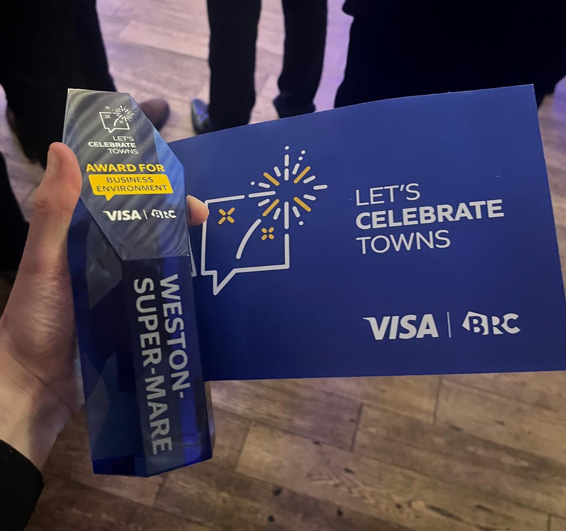 A blue glass award is held next to a blue card with 'Let's Celebrate Towns' printed on it