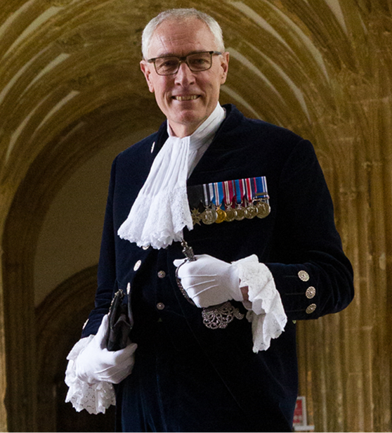 A photo of a white man in glasses with grey hair standing in front of the arches of a church. He is wearing the uniform of the high sheriff with white cravat and gloves, and has medals on his chest. 