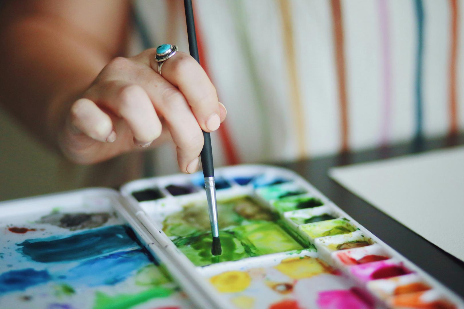 A person dips their paintbrush into green paint on a colourful watercolour palette