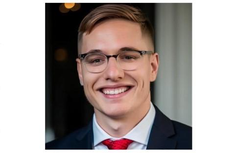 A photograph of a light skinned young man wearing glasses, a white shirt, black suit, and red tie. He is smiling and friendly to the camera.