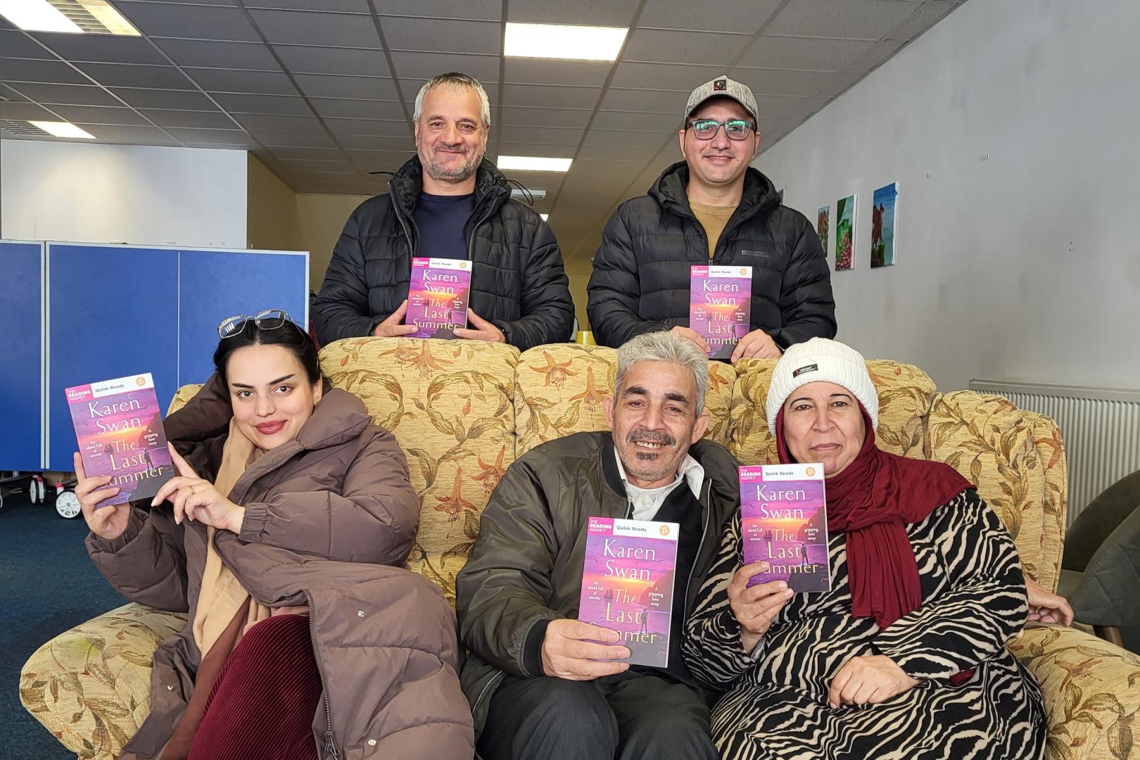 Three people sit on a brown patterned sofa, with two more standing behind them. Everyone is holding a copy of 'The Last Summer', by Karen Swan, and smiling at the camera