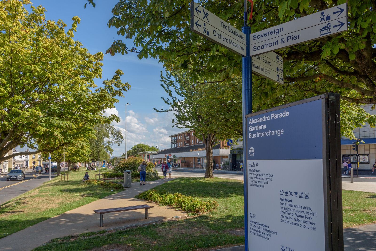 A photograph of a bus hub in Alexandra Parade in Weston. There is a sign to the right showing directions to various town areas, and to the left you can see bus stops through a row of green trees.