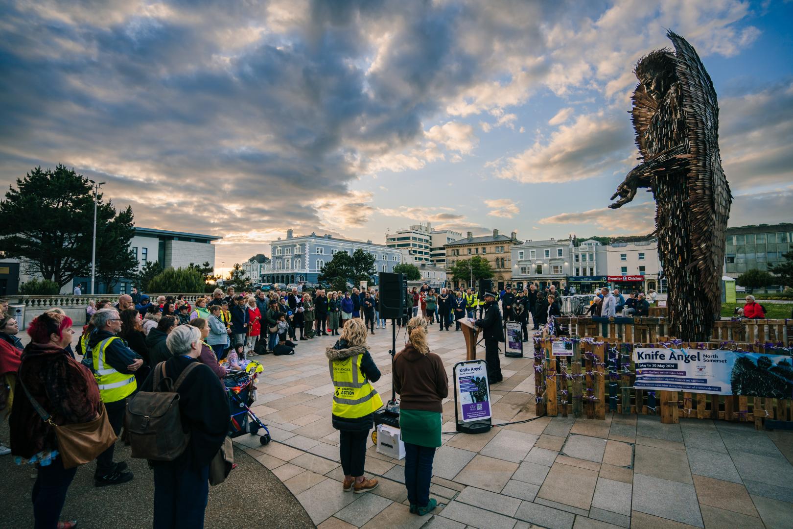 A crowd gathers around the Knife Angel, listening to a police officer speak