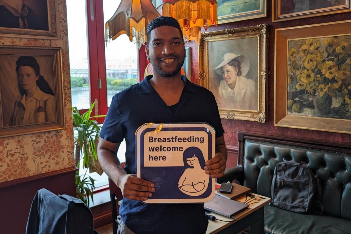 Gagandeep holding a Breastfeeding Welcome sign and smiling at the camera