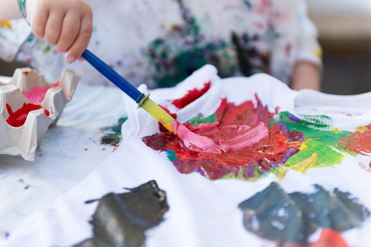 A child uses a paintbrush to swirl colourful paint on a white t-shirt