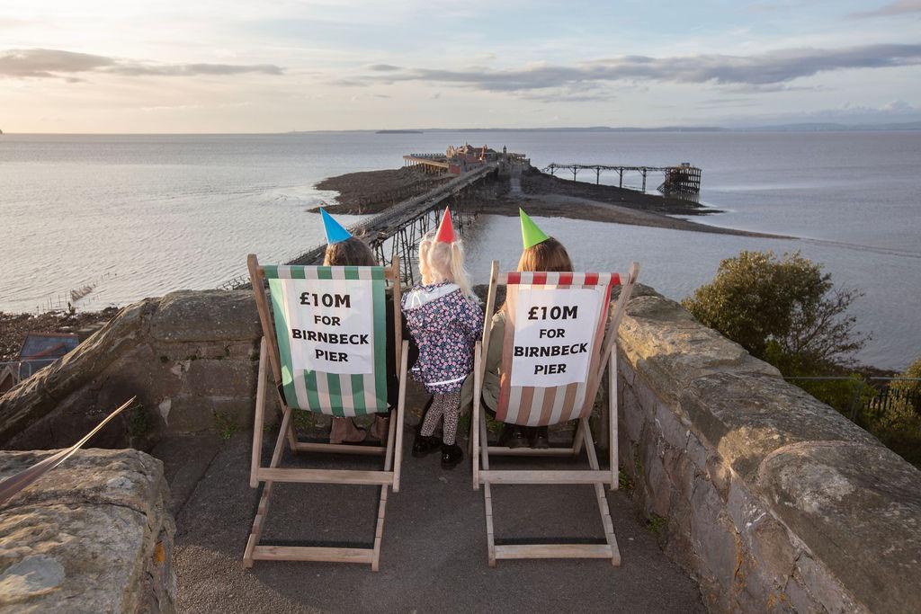 A photo showing two women sitting in traditional wooden deckchairs in Prince Consort Gardens overlooking Birnbeck Pier. A young girl with blonde hair stands between them. All three of them are wearing colourful party hats. Signs on the back of the deckchairs read £10m for Birnbeck Pier.