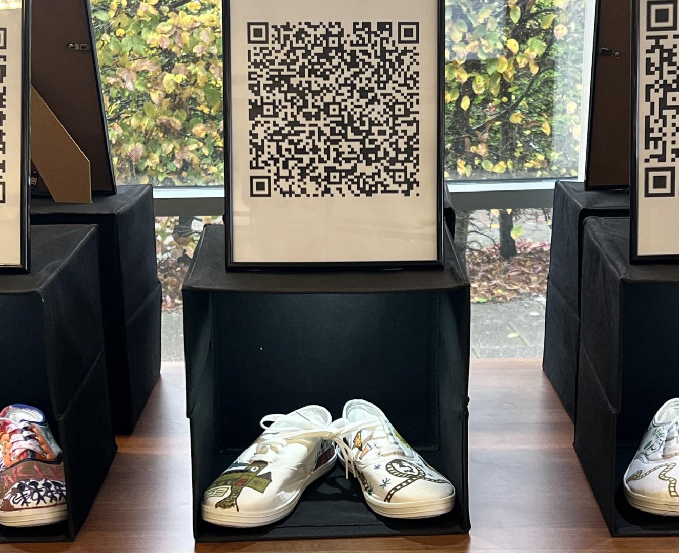 A pair of white trainers decorated with drawings in a black box, with a printed QR code displayed above.