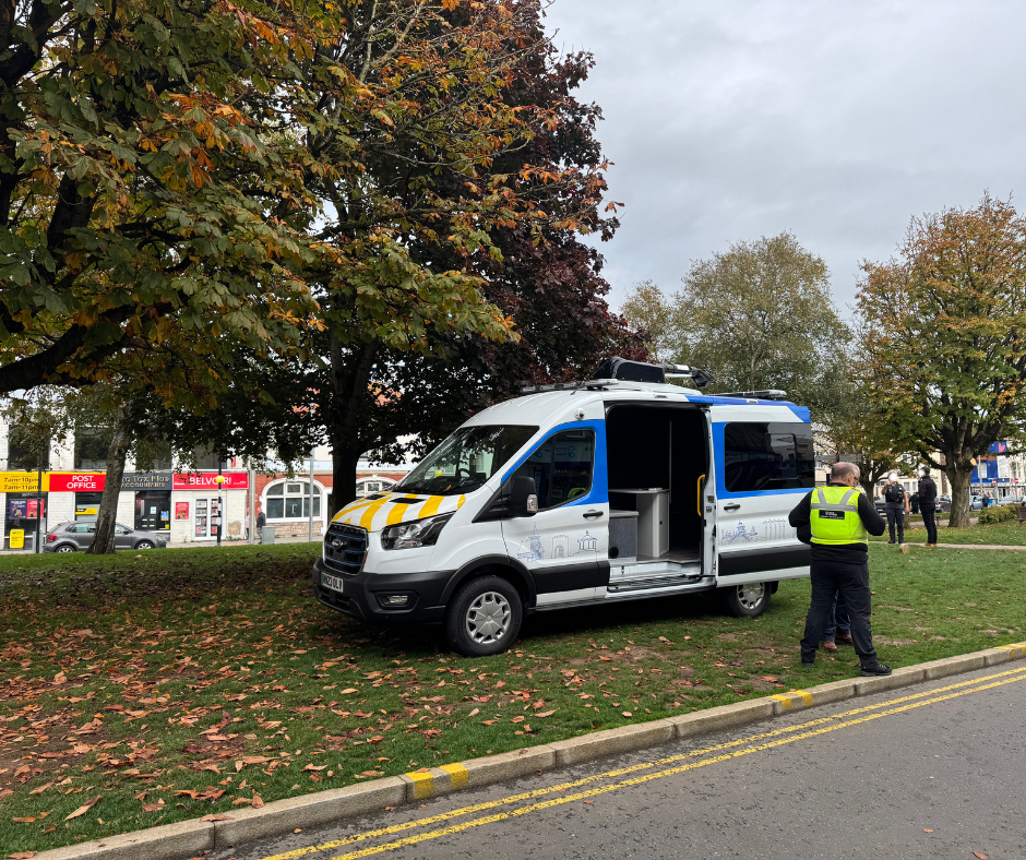 A van is parked on an area of grass with its back doors open, showing the workspace inside. Officials are in discussion nearby