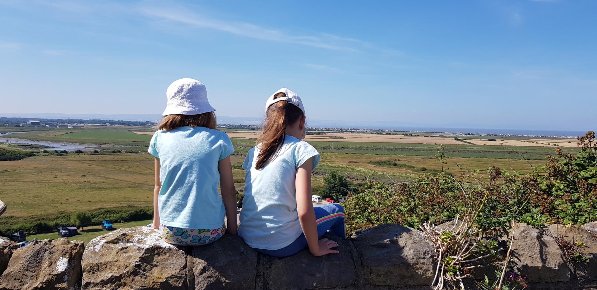 Two children sit on a stone wall, with their backs to the camera. Fields are below them, and the sea is visible in the distance