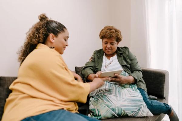 A younger dark skinned woman in a yellow top sits on a couch with an older dark skinned woman with short hair wearing a green jacket. They are sharing an iPad between them and pointing at it. 