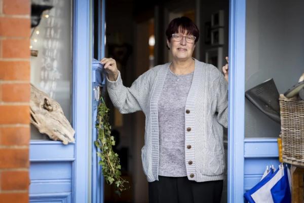 An older white woman with short dark hair and glasses is standing in the doorway of her home, with the doors painted a bright light blue