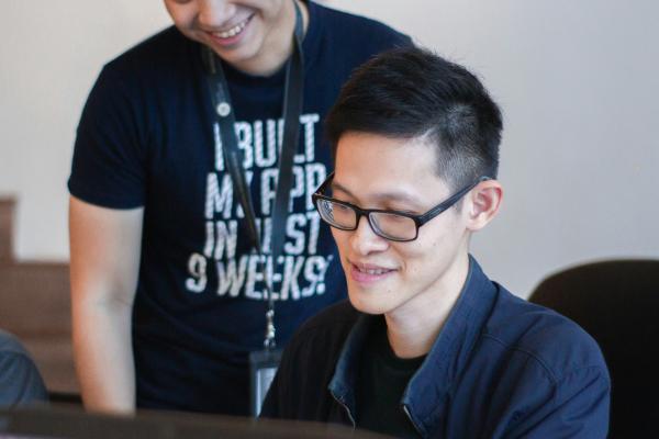 A young Asian man in a black t-shirt with a lanyard around his neck smiles while leaning over to look at a computer screen, there is another Asian man sitting in front of the screen wearing black rimmed glasses and a black buttoned top