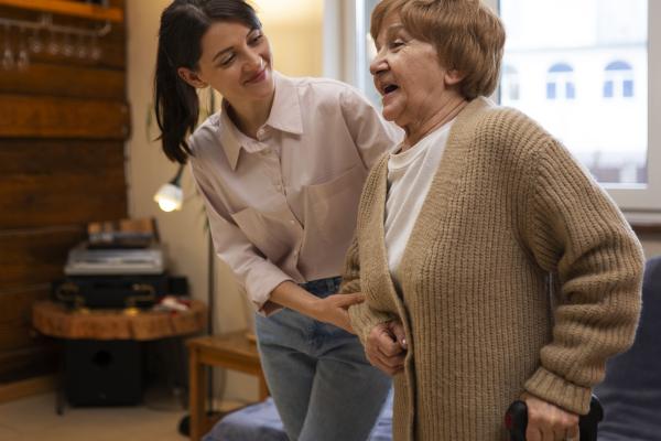 A photograph of two white women, an elderly woman in a long beige cardigan, and a younger woman with dark hair in a ponytail. The younger woman is helping the older woman to walk.